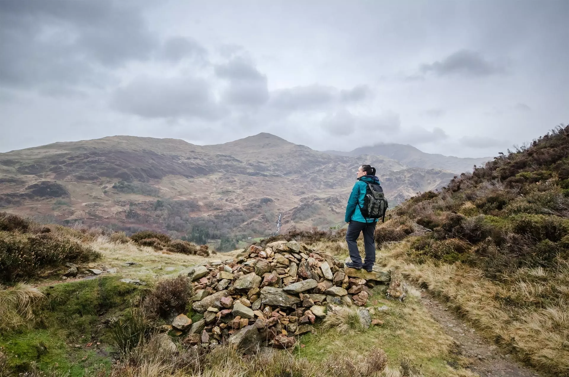 A female hiker wearing a small daypack stands beside a stone trail marker on a rugged path, looking out over the rolling mountain peaks of the Brecon Beacons during a walking holiday in Wales