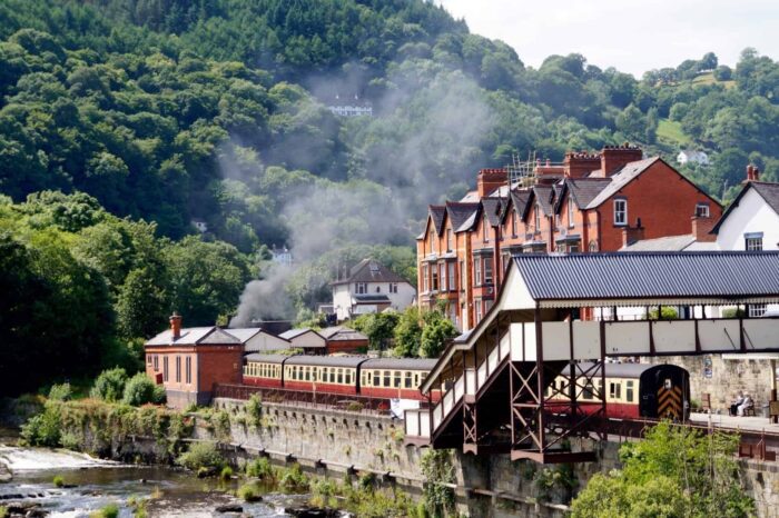 A vintage train passes through a quaint town with red brick buildings, lush green hills, and a riverside railway—one of the best things to do in Wales with children on a bright, sunny day.