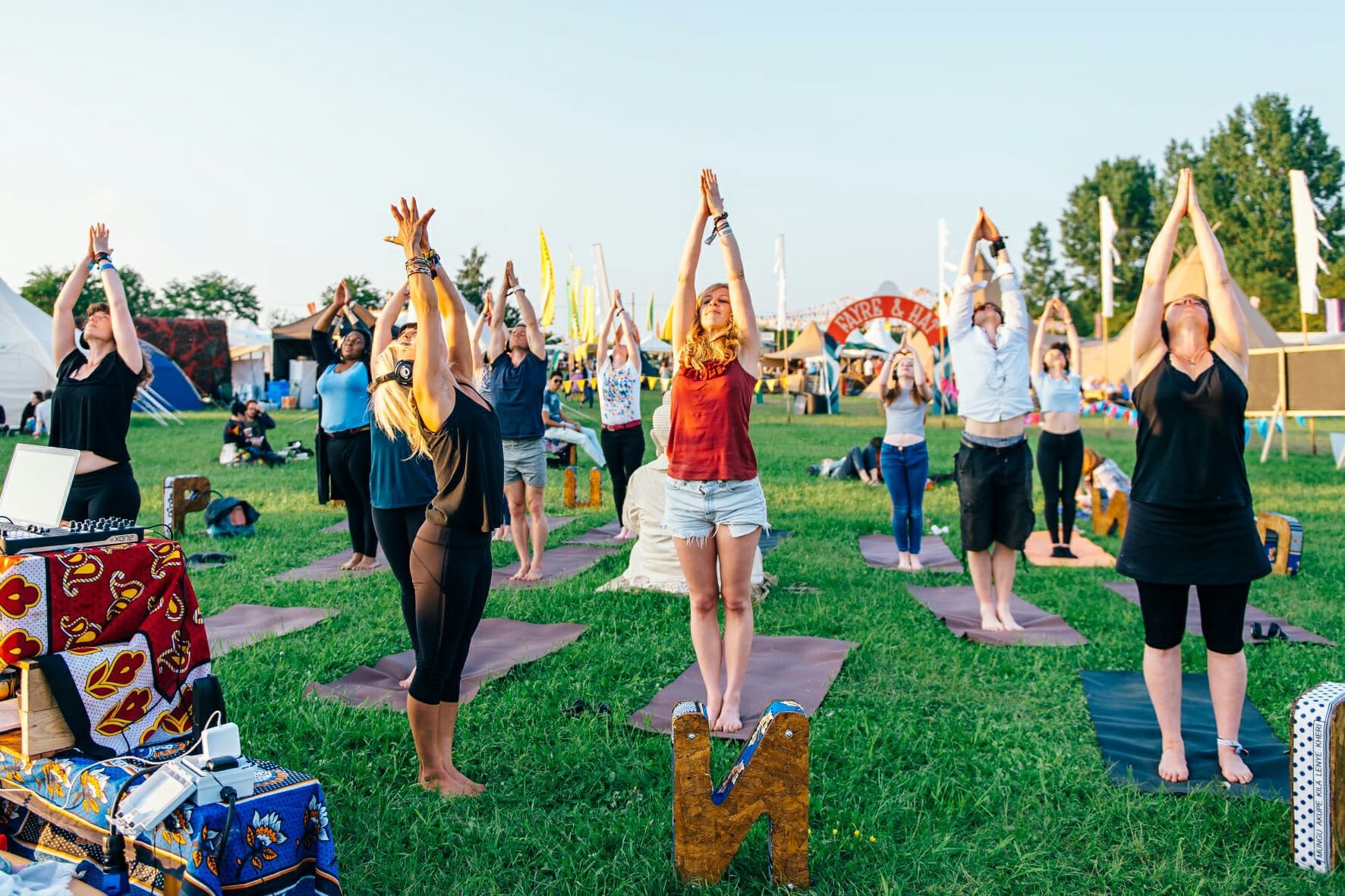 A group of participants practicing yoga outdoors on green grass during a wellness retreat, reaching their arms up towards the sky on colourful mats with trees and tents in the background.