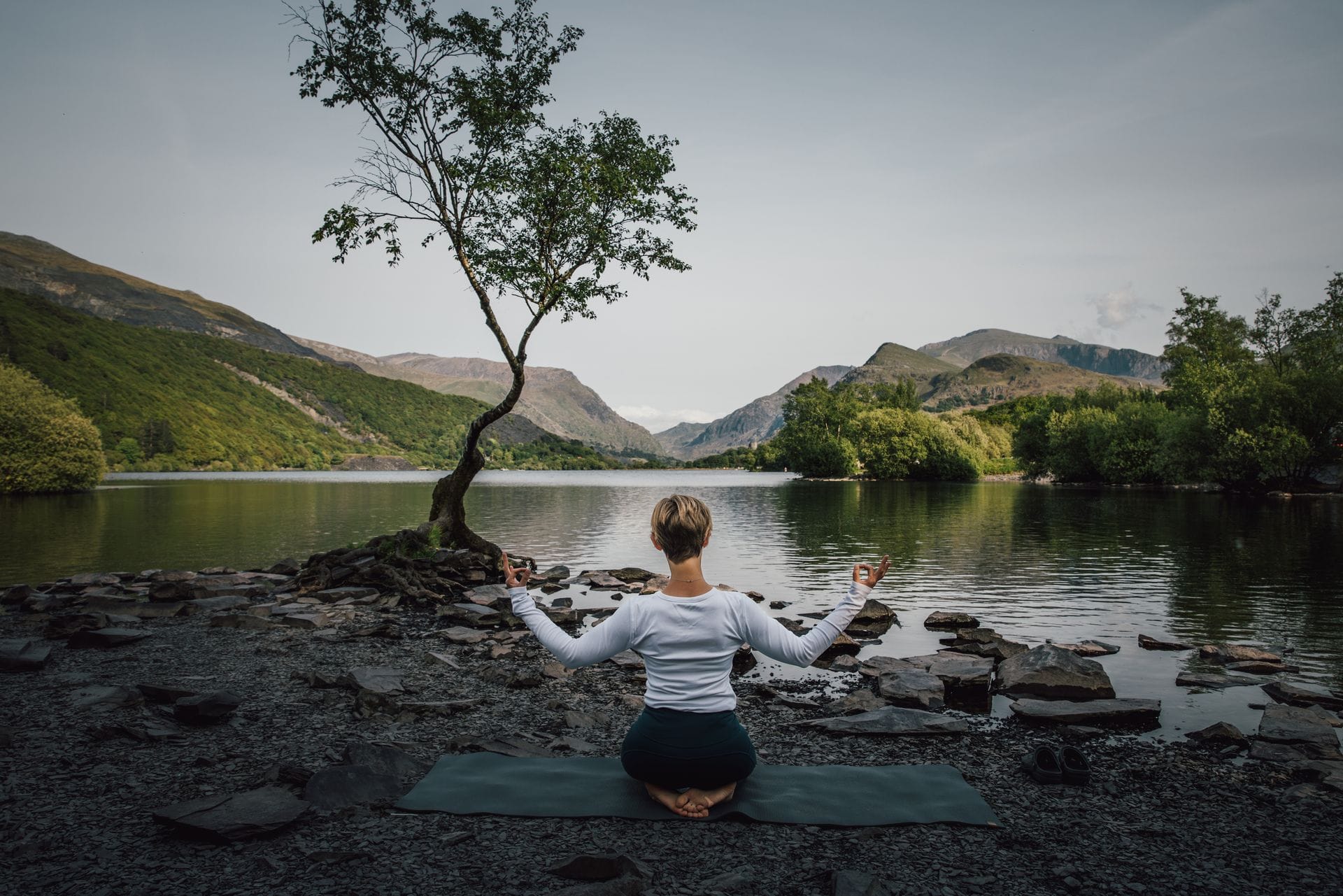A person meditating on a mat beside a calm mountain lake, facing the iconic lone tree on Llyn Padarn during a wellness retreat in Snowdonia